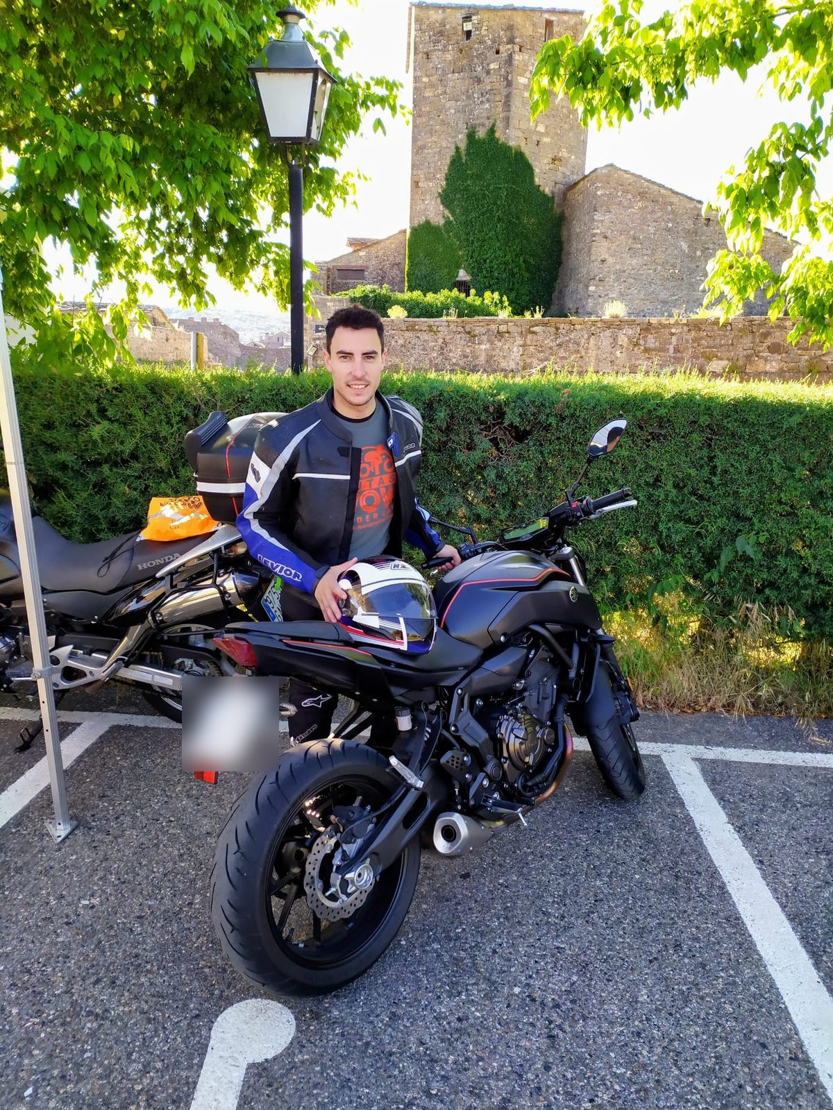 Daniel junto a su moto frente a un pueblo medieval de piedra
